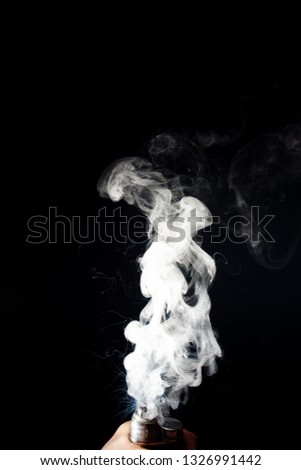 Man hand holds vape. Vape clouds splash from coil on black background. Fog is white. Stock isolated white smoke with spray boiling glycerine around.