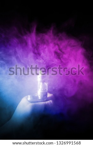 Male hand holds vape in thick vape fog or smoke. Vape clouds around hand with vape on black background. Fog is dual color — blue and purple. Stock isolated colorful smoke with spray glycerine.