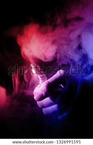 Male hand holds vape with atomiser in thick vape fog or smoke. Vape clouds around hand with vape on black background. Fog is dual color—blue and red. Stock isolated colorful smoke with spray glycerine