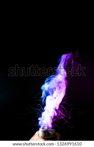Young man holds vape in his hand. Vape clouds splash from vape coil on black background. Fog is dual color — blue and purple. Stock isolated colorful smoke with spray boiling glycerine.