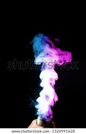 Male hand holds vape. Vape clouds splash from vape coil on black background. Fog is dual color — blue and purple. Stock isolated colorful smoke with spray boiling glycerine.