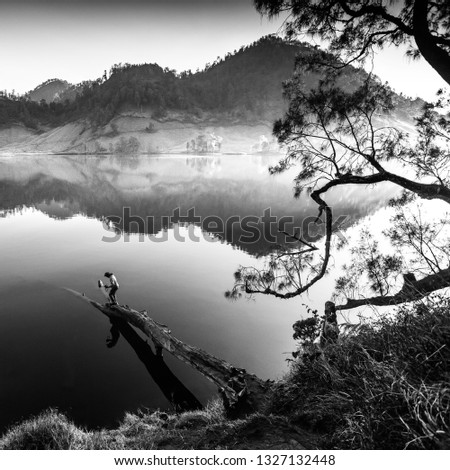 Image of black and white Unidentified hikers stand on dead tree near lake at Ranu Kumbolo,Semeru Volcano Mountain, East Java, Indonesia. Image may contain grain and soft focus.