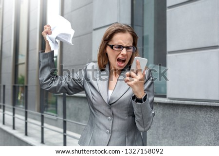 Young businesswoman wearing eyeglasses standing on the city street shouting at busines partner on smartphone angry crumpling the contract