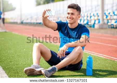 Young sportsman on stadium outside sitting on the field with bottle of water taking selfie photo on smartphone smiling cheerful