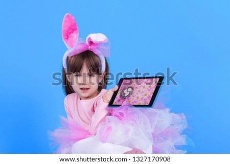 Pink young girl child daughter wears pink dress like rabbit playing in easter holiday game with rabbit ears and tablet with ester eggs on isolated blue background. Spring and Easter holiday concept