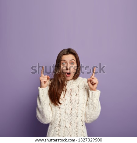 I dont believe what saw. Impressed fearful emotional lady, raises arms and points up, dressed in knitted jumper, impressed by incredible scene, gestures over purple background, empty space above