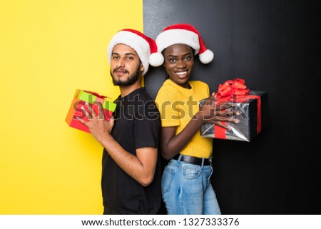 Young mixed race couple, indian man and african woman wearing santa hats with Christmas gift standing isolated on yellow black background
