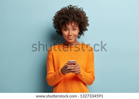 Isolated shot of positive smiling woman holds modern cellular, enjoys wireless internet, checks bank account or sends message in social networks, wears orange clothes, models over blue background