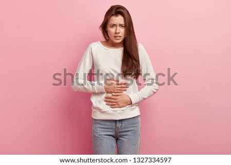 Upset European woman keeps hands on belly, suffers from pain, feels discomfort after eating fast food, wears casual white jumper, isolated over pink background. People, illness, cramps concept Royalty-Free Stock Photo #1327334597