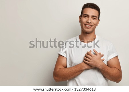 Indoor shot of handsome man keeps hands on chest, being cordial and friendly, expresses gratitude and thankfulness, has pleasant smile, dressed in casual outfit, isolated on white studio wall Royalty-Free Stock Photo #1327334618