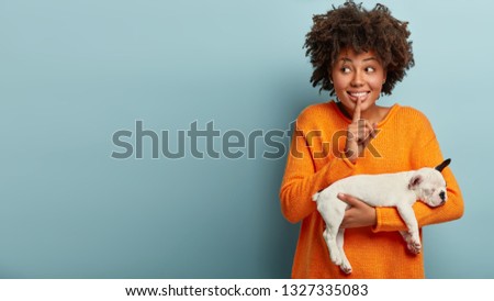 Studio shot of positive dark skinned young female makes shush gesture, looks happily away, holds white and black french bulldog, wears orange jumper, isolated over blue background, free space