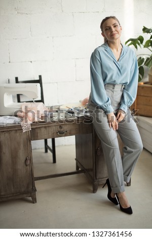 Portrait of confident female tailor, shop owner standing at workplace leaning on desktop.