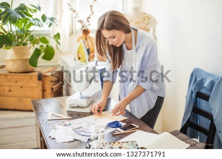 Indoor shot of caucasian young woman in blue casual unbuttoned shirt takes needles for modelling dress from worktable in workshop.