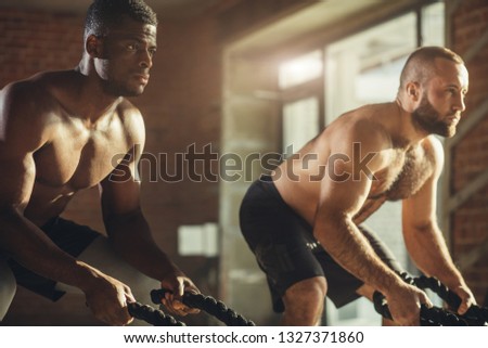 Multiethnic sportive african and caucasian men with bared torso doing double wave during functional training with battle rope in gym, close up. Focus on determined faces of athletes.