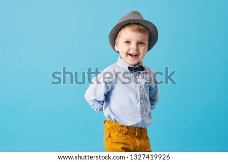 Portrait of happy joyful  little boy isolated on blue background. Toddler child in hat and fashionable suit smiling and have a fun  Royalty-Free Stock Photo #1327419926