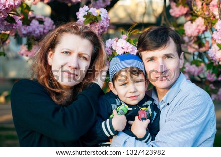 Beautiful woman and her husband hold their son in their hands against the background of pink spring flowers. Familyl love to walk together in warm spring evenings and take pictures
