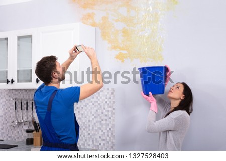 Woman Collecting Water In Blue Bucket From Damaged Ceiling While Repairman Taking Photograph On Mobilephone
