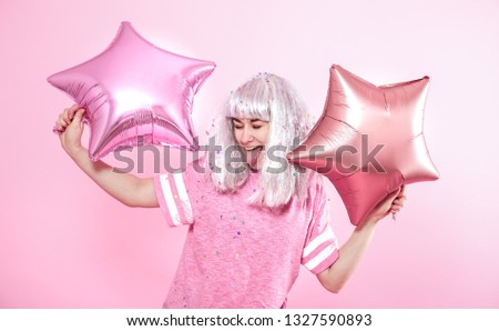 Funny Girl with silver hair gives a smile and emotion on pink background. Young woman or teen girl with balloons and confetti. Concept of holiday and party.