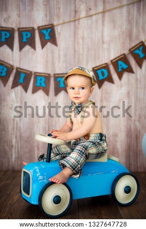 Little boy kid gentleman in retro costume with suspenders and cap is sitting on a wooden car. Children's party with balloons Happy Birthday, 1 year