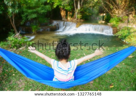 Child or kid girl sit crib or cradle and happy hands up on nature waterfall with green grass and tree in jungle or forest for summer holiday relax or camping picnic and travel trip on family vacation