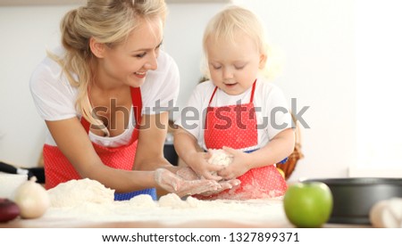 Little girl and her blonde mom in red aprons  playing and laughing while kneading the dough in kitchen. Homemade pastry for bread, pizza or bake cookies. Family fun and cooking concept