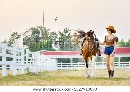 Active Asia cowgirl in hat walking with horse. Farmer girl in countryside ranch. Beautiful woman training and taking care of her horse with love and caring.
