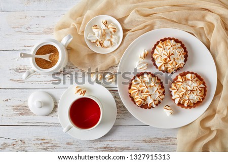 close-up of Lemon meringue mini pies with browned meringue peaks served with tea, mini marshmallows and brown organic sugar on wooden table with light cloth, view from above