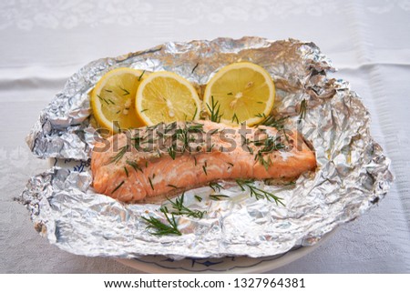 Close up picture salmon fillet baked in aluminium baking foil, low fat, fresh, healty dish served as a lunch or dinner in garden restaurant on white plate and slices of organic lemon and chopped dill.