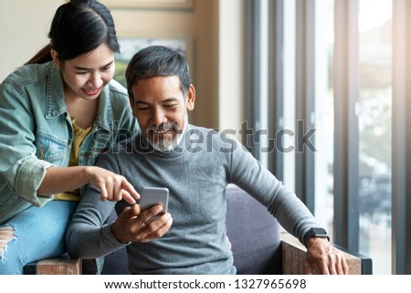 Attractive mature asian man with white stylish short beard looking at smartphone computer with teenage eye glasses hipster woman in cafe. Teaching internet online or wifi technology concept Royalty-Free Stock Photo #1327965698
