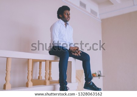 Lifestyle portrait of handsome young black man sitting at parapet at his decorated in african style room with plants near wall and stairs. Indoor portrait of happy dark-skinned nigerian male resting.