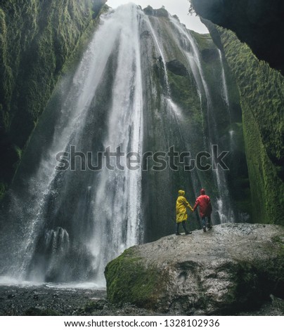 Couple in raincoats looking on scenic high waterfall. Beautiful waterfall in Iceland
