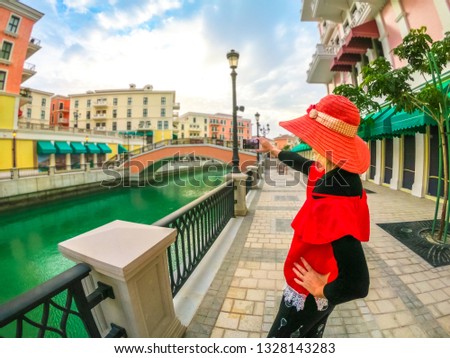 Woman in sunhat takes photo with smartphone of Venice, Venetian style waterfront village. Tourism in Middle East. Photographer takes pictures from Qanat Quartier in the Pearl-Qatar, Persian Gulf.
