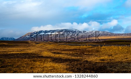 Photo describes a typical Icelandic landscape near Reykjavik. Orange and brown meadows with shadows from clouds and snowy mountain in the background. Turquise sky with smoky clouds tht is Icleland