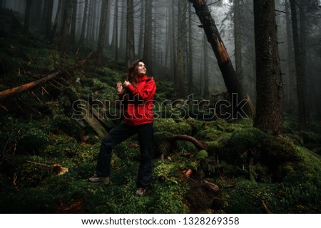 Young woman traveller in red jacket in the misty landscape with mossy rocks in fir forest. Fog, rain and clouds in the mountains. Adventure travel in wildlife nature