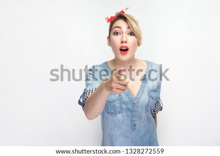Wow, its unbelievable. Portrait of shocked beautiful young woman in casual blue shirt with makeup and red headband standing and pointing at camera. indoor studio shot, isolated on white background.