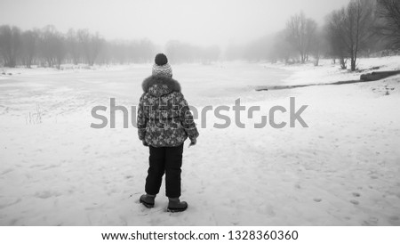 Winter day. The girl on the river bank looks at the lake. Fog and light rain with snow. Picture taken in Ukraine, Kiev region. Horizontal frame. Black and white image