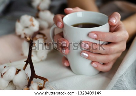 Female hands with pastel colored manicure holding a white mug with beverage on bright background decorated with cotton flowers. Close up. Blurred background