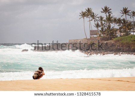 Young man on north shore beach of Oahu, Hawaii taking travel landscape pictures.