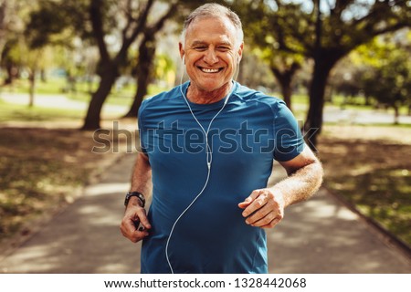 Portrait of a senior man in fitness wear running in a park. Close up of a smiling man running while listening to music using earphones. Royalty-Free Stock Photo #1328442068