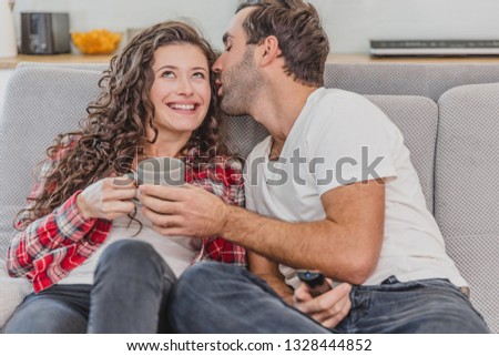 Beautiful hilarious young man holding a remote control. During this, she watches a whirlwind and kisses her cheek. Put two on the couch at home.