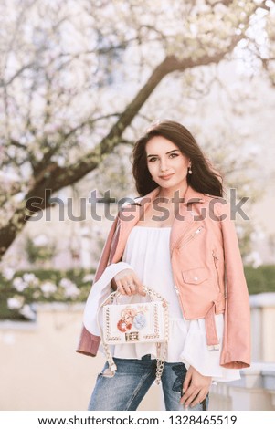 Outdoor portrait of young beautiful fashionable lady wearing pink leather jacket, holding small stylish white bag with floral application. Blooming tree on background. Spring fashion concept