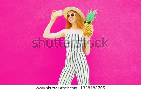 Stylish happy smiling woman with pineapple taking selfie picture by phone in summer round hat, white striped jumpsuit on colorful pink background