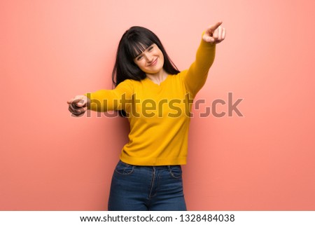 Woman with yellow sweater over pink wall points finger at you while smiling