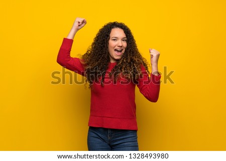 Teenager girl with red sweater over yellow wall celebrating a victory
