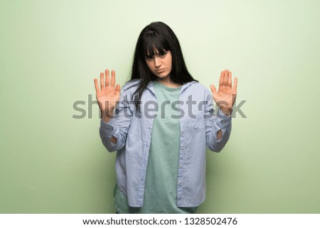 Young woman over green wall making stop gesture with both hands