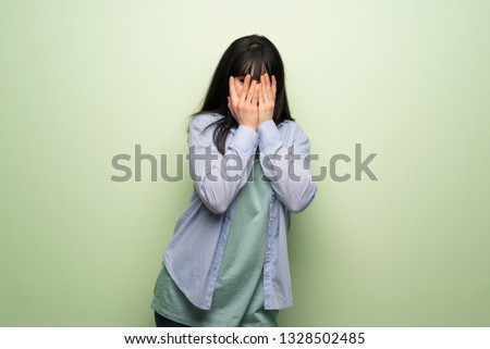 Young woman over green wall covering eyes by hands and looking through the fingers
