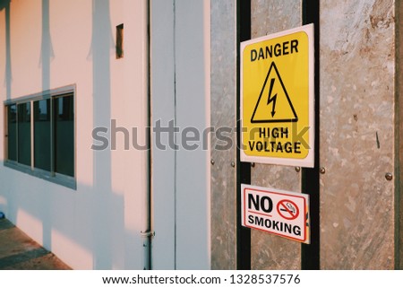 Warning Danger high Voltage Sign and no smoking Label on steel pipe cover with sunlight and Electric pole's shadow on surface of white wall, Safety sign and symbol concept 