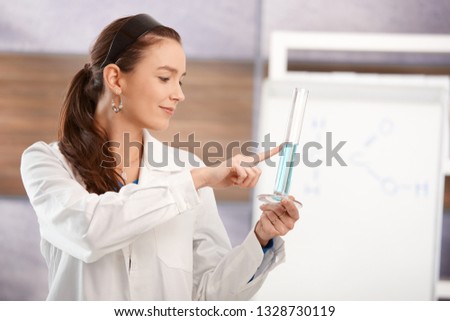 Young female teacher teaching chemistry in school classroom, looking at test tube, smiling.