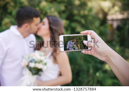Someone is taking a picture on the phone bride and groom while they kiss. Beautiful couple in white