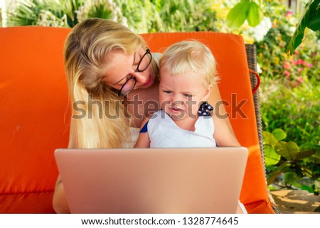 fair-haired one year old female kid looking at a laptop cartoon with mother blonde freelancer surfer on sunbeds resort summer beach.business woman freelancing and daughter relaxing by the sea